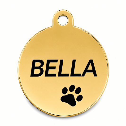A round gold-colored metal pet ID tag with a hanging loop at the top, featuring the name 'BELLA' engraved in bold black letters and a black paw print below it, shown on a clean white background.