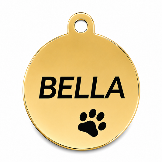 A round gold-colored metal pet ID tag with a hanging loop at the top, featuring the name 'BELLA' engraved in bold black letters and a black paw print below it, shown on a clean white background.